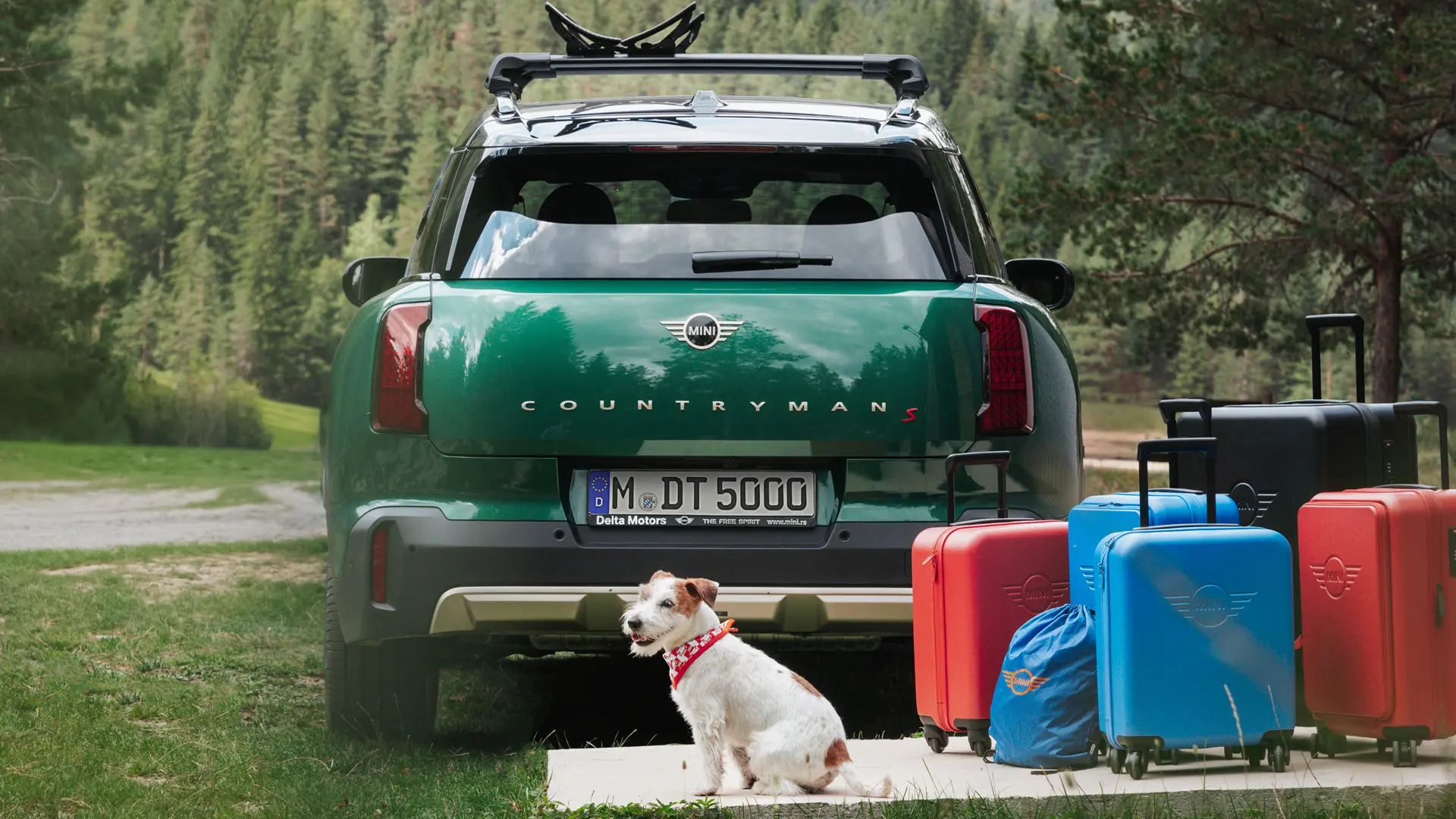 MINI Countryman in green with various luggage items available from the MINI Lifestyle store standing behind the vehicle together with a Jack Russell dog. 